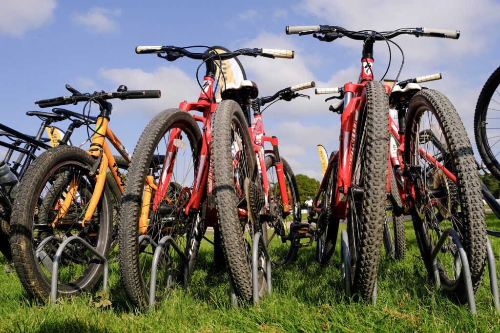 A row of bikes lean in a stand on grass with a blue sky.