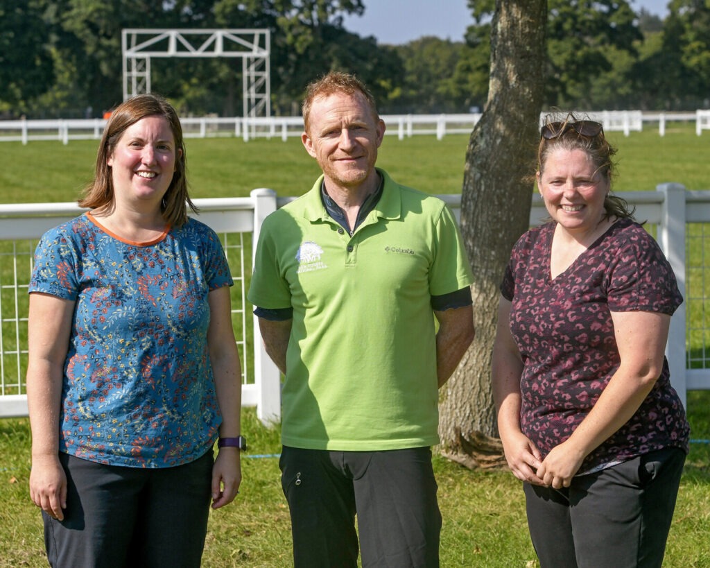 Three people stand in a field - two women with a man between them.