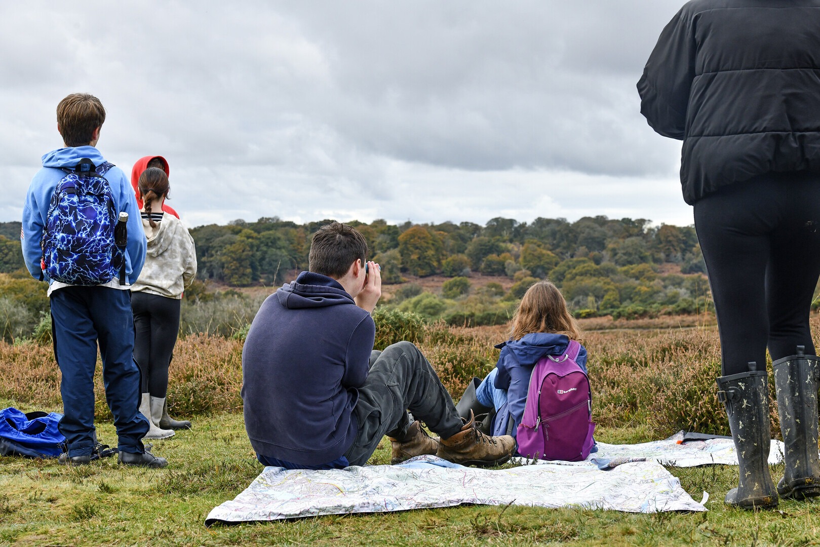 A group of young people look out over heathland. Two are sat on a mat and two are standing.
