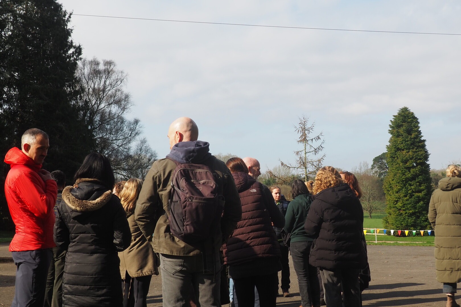 A group of people stand at the edge of a green field listening to a talk with their back to the camera.