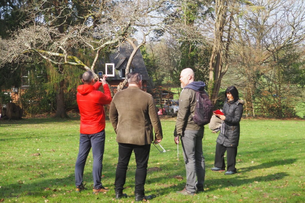 Four people stand in a field with trees during a nature connection session. One is looking through a photo frame with his phone camera out.