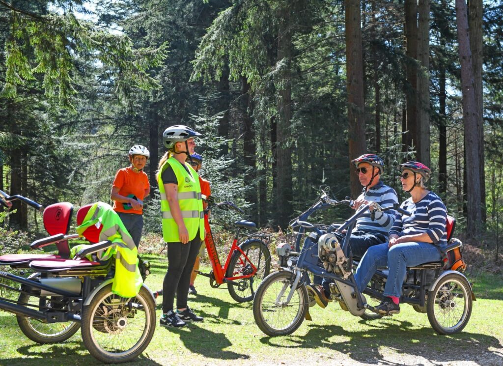 Cyclists in helmets talk in a wooded park beside a three-wheeled tandem bike with two older riders seated side by side